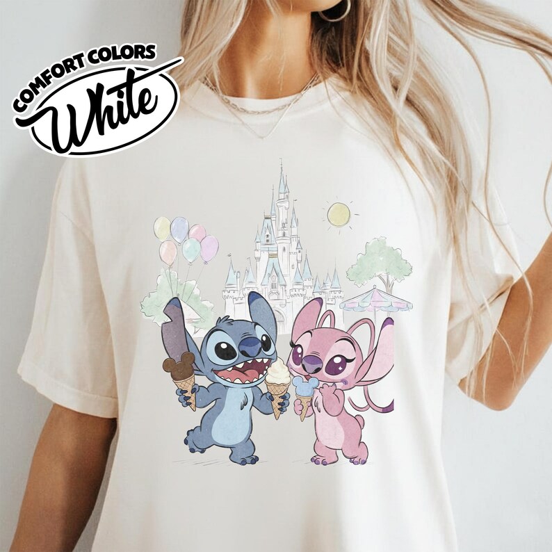 Puede incluir: Camiseta blanca Comfort Colors con una ilustraci&oacute;n de dibujos animados de Stitch y Angel sosteniendo conos de helado frente a un castillo. La camiseta tambi&eacute;n tiene un gr&aacute;fico de globos, un sol y un &aacute;rbol.