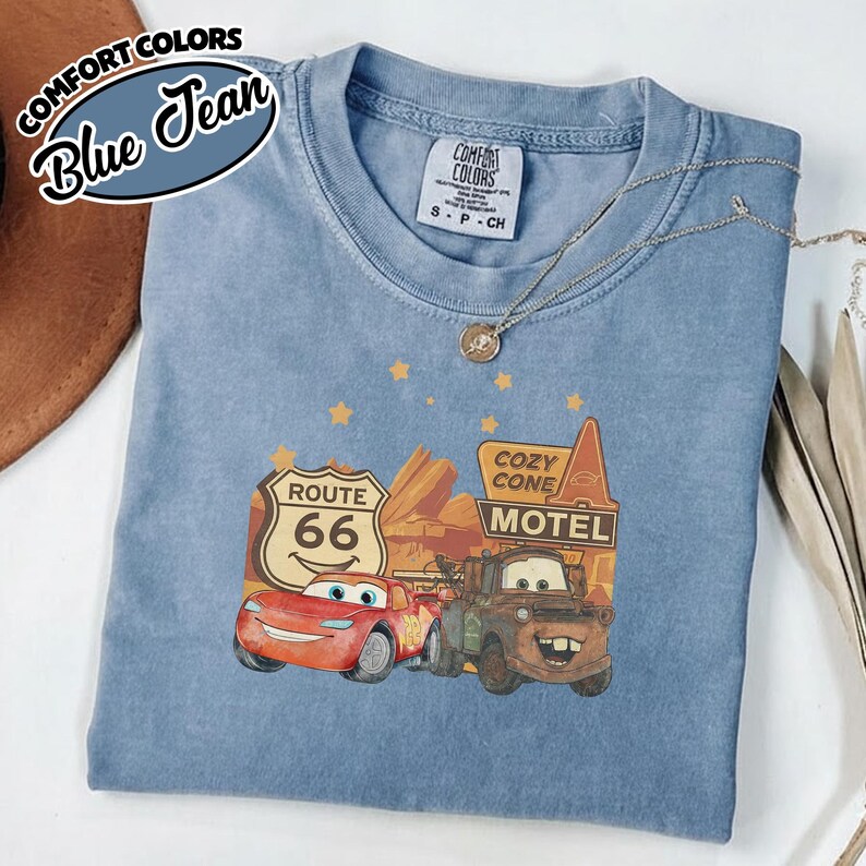 May include: Blue jean Comfort Colors t-shirt featuring a graphic of Lightning McQueen and Mater from the movie Cars, with Route 66 and Cozy Cone Motel signage. The shirt has a crew neck and short sleeves.