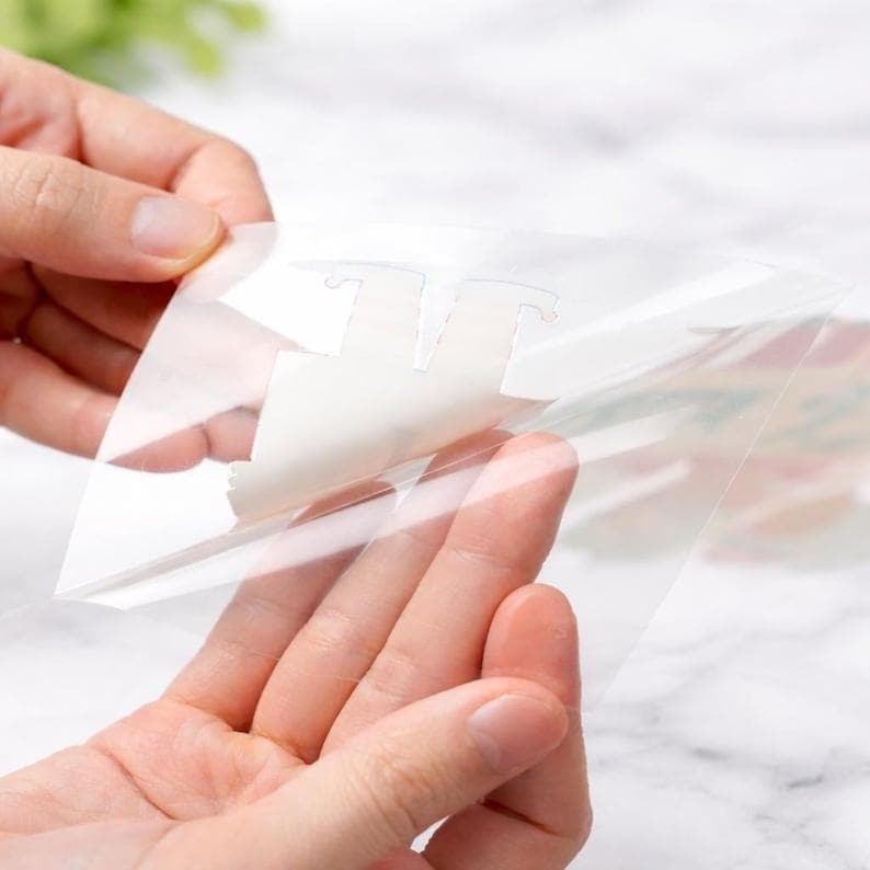 May include: A clear adhesive transfer sheet being peeled back to reveal a white die-cut shape underneath. The transparent film is flexible and smooth, showing a portion of the white material separating from the backing as it is lifted.