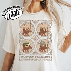 May include: White Comfort Colors t-shirt with a graphic of four cartoon characters holding spears, each in a circular frame. The text "FEAR THE KAKAMORA" is printed below the graphic. The shirt also has the text "Comfort Colors White" in a circular design.