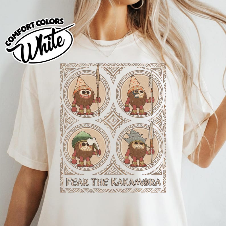 May include: White Comfort Colors t-shirt with a graphic of four cartoon characters holding spears, each in a circular frame. The text "FEAR THE KAKAMORA" is printed below the graphic. The shirt also has the text "Comfort Colors White" in a circular design.