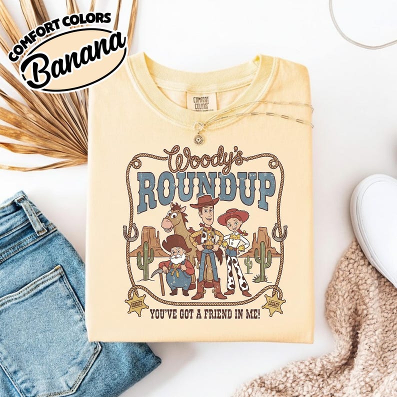 May include: A light yellow t-shirt with the text "Woody's Roundup" and a graphic of characters from the movie Toy Story. The shirt also has the text "You've Got a Friend in Me!" and the words "Comfort Colors" and "Banana".