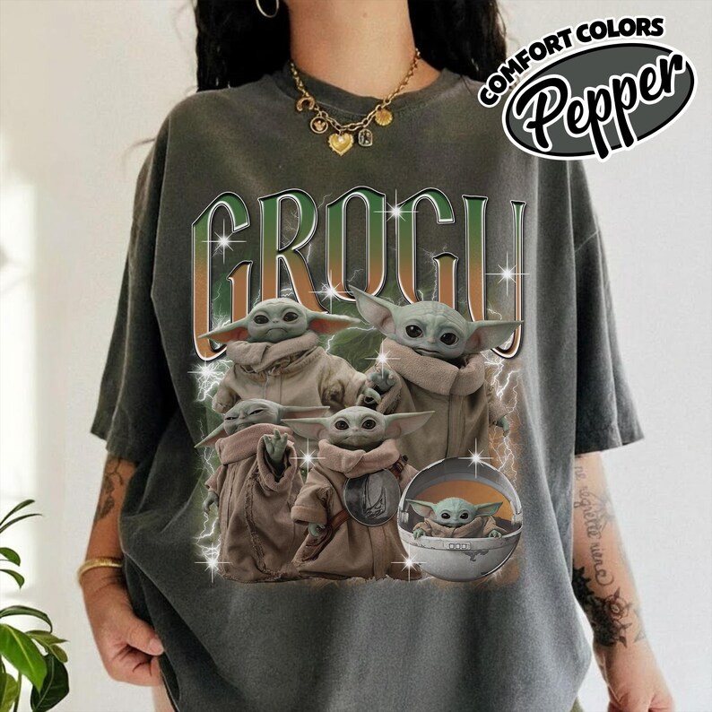 K&ouml;nnte beinhalten: Ein anthrazitfarbenes Kurzarm-T-Shirt mit einem Grafikdruck mehrerer Grogu-Figuren in beigen Gew&auml;ndern. Der Text GROGU erscheint in gro&szlig;en gr&uuml;nen und orangefarbenen Buchstaben &uuml;ber den Figuren, akzentuiert durch wei&szlig;e Blitze und funkelnde Sterne.