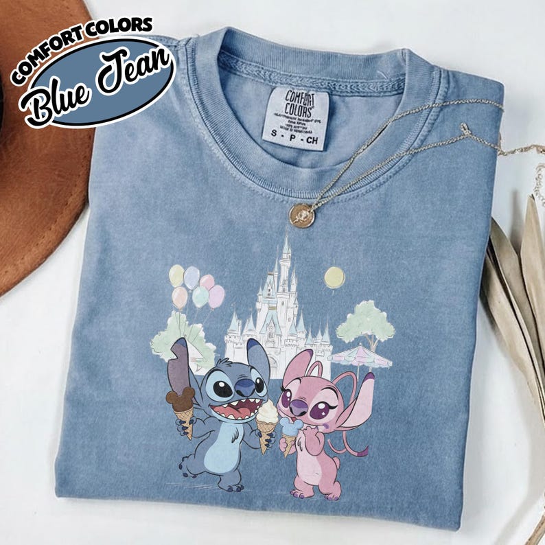 Puede incluir: Camiseta Comfort Colors azul vaquero con un gr&aacute;fico de Stitch y Angel de Lilo & Stitch, sosteniendo conos de helado frente a un castillo. La camiseta tiene cuello redondo y mangas cortas. El texto "Comfort Colors" y "Blue Jean" est&aacute; arriba a la izquierda.
