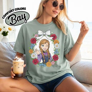 Peut inclure: T-shirt vert clair avec un personnage de dessin anim&eacute; dans une couronne florale. Le personnage a des tresses blondes et fait un clin d'&oelig;il et fait signe de la main. La couronne comprend des roses rouges, des marguerites blanches et d'autres fleurs. Le t-shirt porte l'inscription "Comfort Colors Bay".