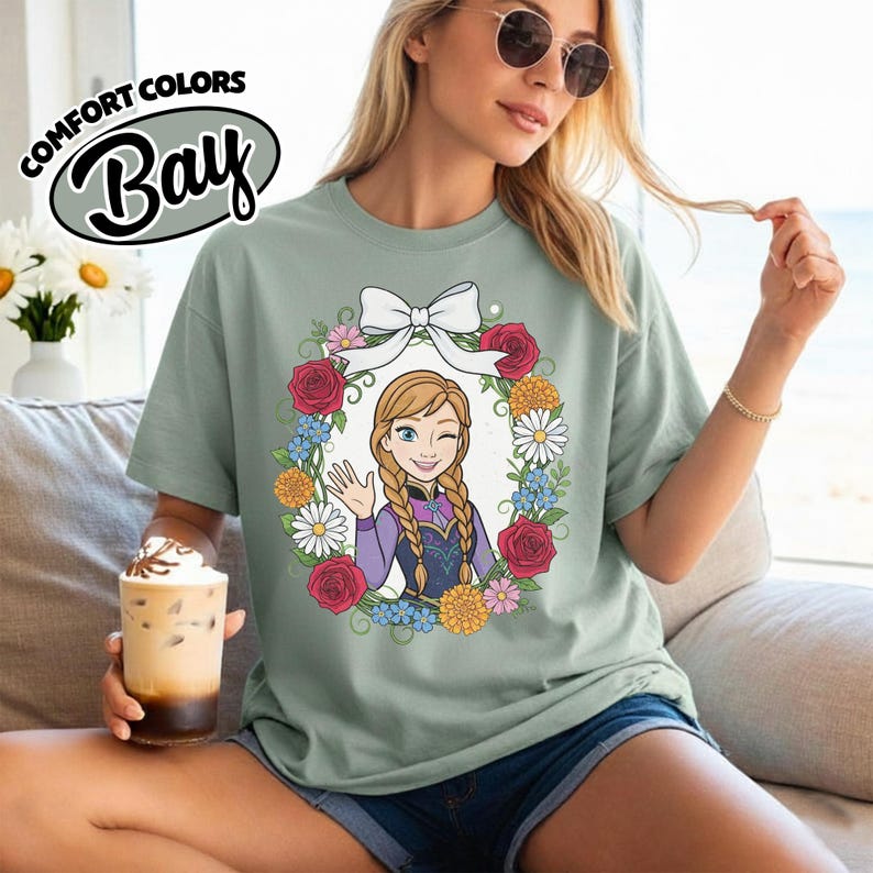 Peut inclure: T-shirt vert clair avec un personnage de dessin anim&eacute; dans une couronne florale. Le personnage a des tresses blondes et fait un clin d'&oelig;il et fait signe de la main. La couronne comprend des roses rouges, des marguerites blanches et d'autres fleurs. Le t-shirt porte l'inscription "Comfort Colors Bay".