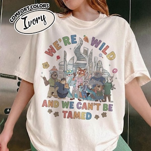 May include: Ivory-colored t-shirt with a colorful graphic featuring cartoon animal characters and the text "WE'RE WILD AND WE CAN'T BE TAMED." The shirt also has the words "COMFORT COLORS" and "Ivory" in a circle.