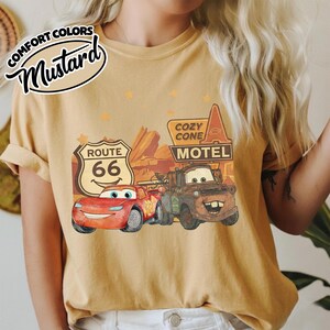 May include: Mustard-colored t-shirt featuring a graphic of the Cars movie characters, Lightning McQueen and Mater, with a Route 66 sign and Cozy Cone Motel. The shirt has the words "Comfort Colors" and "Mustard" in a circular design.