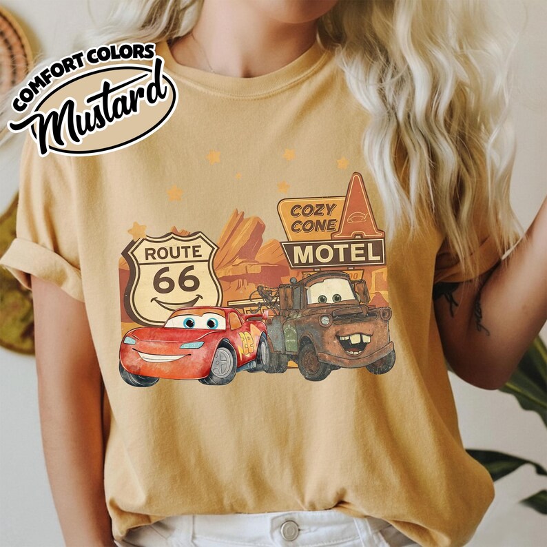May include: Mustard-colored t-shirt featuring a graphic of the Cars movie characters, Lightning McQueen and Mater, with a Route 66 sign and Cozy Cone Motel. The shirt has the words "Comfort Colors" and "Mustard" in a circular design.