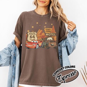 May include: Espresso-colored t-shirt featuring a graphic of characters from the movie Cars, including Lightning McQueen and Mater, with Route 66 and Cozy Cone Motel signs. The shirt has a vintage, distressed look.