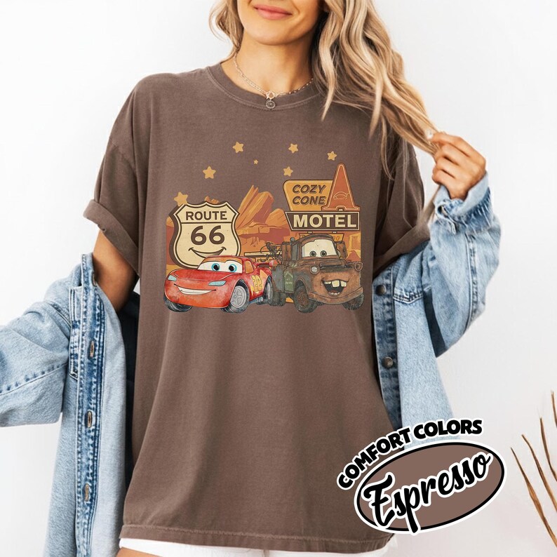 May include: Espresso-colored t-shirt featuring a graphic of characters from the movie Cars, including Lightning McQueen and Mater, with Route 66 and Cozy Cone Motel signs. The shirt has a vintage, distressed look.