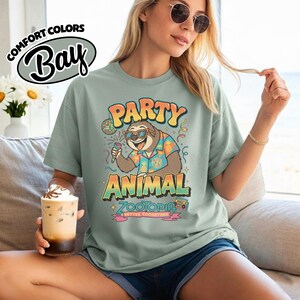 Puede incluir: Una camiseta verde claro con el texto "PARTY ANIMAL" y un perezoso de dibujos animados con gafas de sol y una camisa hawaiana. La camiseta tambi&eacute;n tiene el texto "ZOOTOPIA" y "BETTER ZOOGETHER".