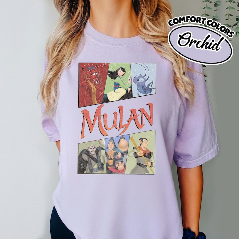 Puede incluir: Una camiseta de color lila claro con un gr&aacute;fico que presenta personajes de la pel&iacute;cula animada Mulan. El dise&ntilde;o incluye la palabra "Mulan" en letras naranjas y varias ilustraciones de personajes. La camiseta tambi&eacute;n tiene las palabras "Comfort Colors" y "Orchid".