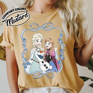 May include: Mustard-colored t-shirt featuring a cartoon image of Elsa, Anna, and Olaf from Frozen, framed by a blue floral design. The text "COMFORT COLORS" and "Mustard" are printed on the top left of the shirt.