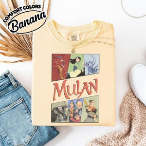 Puede incluir: Camiseta gr&aacute;fica de Mulan de color amarillo claro. La camiseta presenta un dise&ntilde;o de c&oacute;mic con personajes de la pel&iacute;cula y la palabra "Mulan" en rojo. La camiseta tiene las palabras "Comfort Colors" y "Banana".