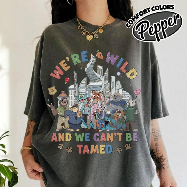 May include: Dark gray t-shirt with a colorful graphic featuring characters and text from the movie "Zootopia." The text reads "WE'RE WILD AND WE CAN'T BE TAMED." The shirt has a relaxed fit and short sleeves.