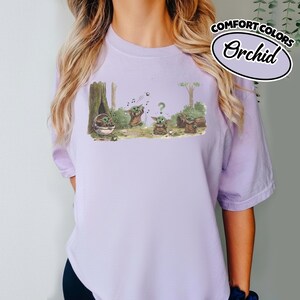 May include: A light purple Comfort Colors t-shirt with a whimsical illustration of a character in a forest setting. The graphic features the character in various poses, with musical notes and a question mark. The text "Comfort Colors Orchid" is printed on the upper right.