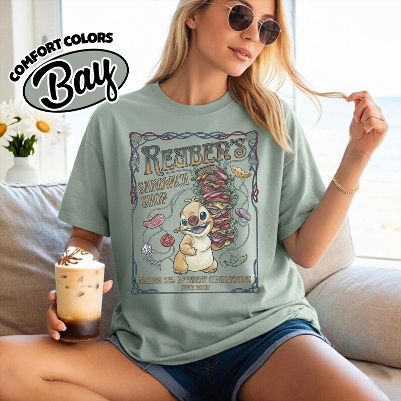 K&ouml;nnte beinhalten: Hellgr&uuml;nes T-Shirt mit einer Grafik eines Zeichentrickcharakters, der ein gro&szlig;es Sandwich h&auml;lt. Die Grafik zeigt "Reuber's Sandwich Shop" und "Making 625 Different Condiments Since 2001". Das Shirt hat auch die Worte "Comfort Colors Bay".