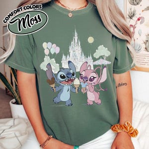 Puede incluir: Camiseta verde con un dise&ntilde;o de Stitch y Angel sosteniendo conos de helado frente a un castillo. La parte superior izquierda muestra "Comfort Colors" y "Moss" en un dise&ntilde;o circular. El dise&ntilde;o est&aacute; impreso en una camiseta verde.