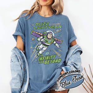 May include: A blue jean colored t-shirt featuring a graphic of Buzz Lightyear flying with wings extended. The text reads BUZZ LIGHTYEAR TO INFINITY AND BEYOND. The design includes planets, stars, and comets in a space theme on the front of the shirt.