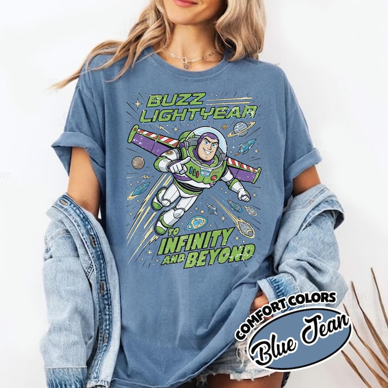 May include: A blue jean colored t-shirt featuring a graphic of Buzz Lightyear flying with wings extended. The text reads BUZZ LIGHTYEAR TO INFINITY AND BEYOND. The design includes planets, stars, and comets in a space theme on the front of the shirt.