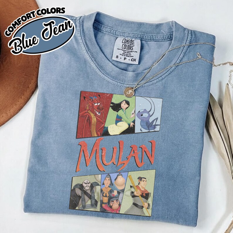 Puede incluir: Una camiseta azul con un gr&aacute;fico de personajes de la pel&iacute;cula Mulan. La palabra "Mulan" est&aacute; impresa en naranja. La esquina superior izquierda de la camiseta tiene las palabras "Comfort Colors" y "Blue Jean". Un collar plateado est&aacute; sobre la camiseta.