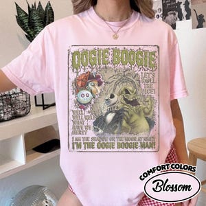 May include: A light pink Comfort Colors t-shirt featuring a graphic of Oogie Boogie from The Nightmare Before Christmas. The graphic includes text that reads "Oogie Boogie" and "I'm the Oogie Boogie Man!"