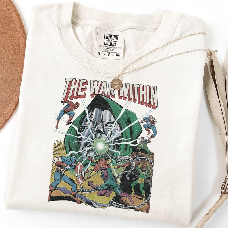 May include: Cream-colored t-shirt featuring a comic book-style graphic with the text "THE WAR WITHIN." The graphic depicts various superheroes battling a villain in a green cloak. A gold necklace with a pendant is draped over the shirt.