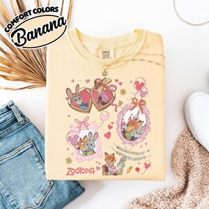 May include: A pale yellow t-shirt featuring a graphic print of characters from Zootopia, including Judy Hopps and Nick Wilde in heart-shaped and ornate frames, surrounded by small hearts, balloons, and the Zootopia logo in pink text.