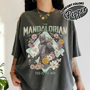 May include: A charcoal gray short-sleeve t-shirt featuring a graphic of The Mandalorian and Grogu surrounded by colorful flowers. The text reads THE MANDALORIAN and THIS IS THE WAY. The shirt has a vintage-style wash and a relaxed, loose fit.