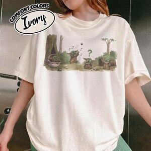May include: Ivory-colored t-shirt with a cartoon graphic of a character from a popular science fiction series. The graphic depicts the character in various poses in a forest setting. The shirt has the words "COMFORT COLORS" and "Ivory" printed on the top left.