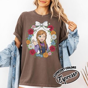 Peut inclure: T-shirt marron avec une illustration de dessin anim&eacute; d'un personnage aux cheveux tress&eacute;s, faisant un clin d'&oelig;il et saluant, entour&eacute; d'une couronne florale et d'un n&oelig;ud blanc. Le texte "COMFORT COLORS Espresso" est imprim&eacute; en bas &agrave; droite.