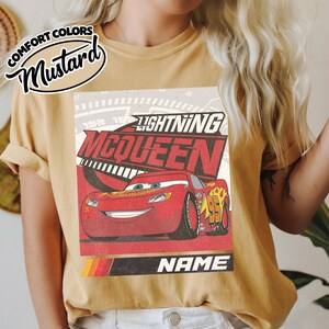 May include: A mustard yellow t-shirt featuring a graphic print of the red race car Lightning McQueen. The design includes the text Lightning McQueen, a speedometer background, and a black bar at the bottom with the word NAME in white.