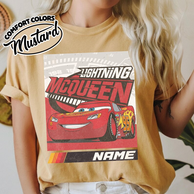 May include: A mustard yellow t-shirt featuring a graphic print of the red race car Lightning McQueen. The design includes the text Lightning McQueen, a speedometer background, and a black bar at the bottom with the word NAME in white.