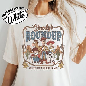 May include: White Comfort Colors t-shirt featuring a graphic of Woody's Roundup characters. The design includes Woody, Jessie, and other characters with the text "Woody's Roundup" and "You've Got a Friend in Me!"