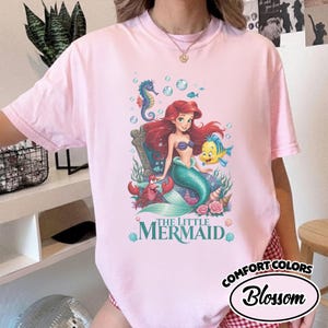 May include: Pink t-shirt featuring a colorful illustration of Ariel from The Little Mermaid. The graphic includes Ariel, a seahorse, a crab, and a yellow fish. The text "THE LITTLE MERMAID" is below the illustration. The shirt is labeled "Comfort Colors Blossom".