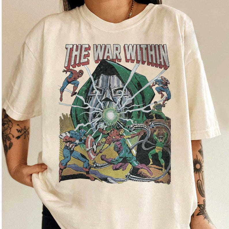 May include: Cream-colored t-shirt featuring a comic book-style graphic with the text "THE WAR WITHIN." The graphic depicts several superheroes, including Spider-Man and Captain America, in a battle scene with a central villain.