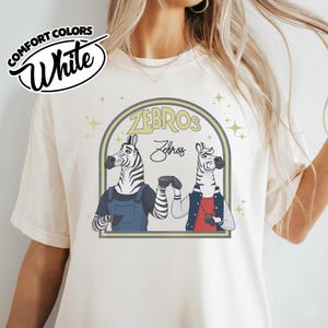 May include: Cream-coloured t-shirt with a graphic of two zebras in a stylized design. The zebras are depicted in a friendly pose, with the word "ZEBROS" and "Zebros" in a decorative font above them.