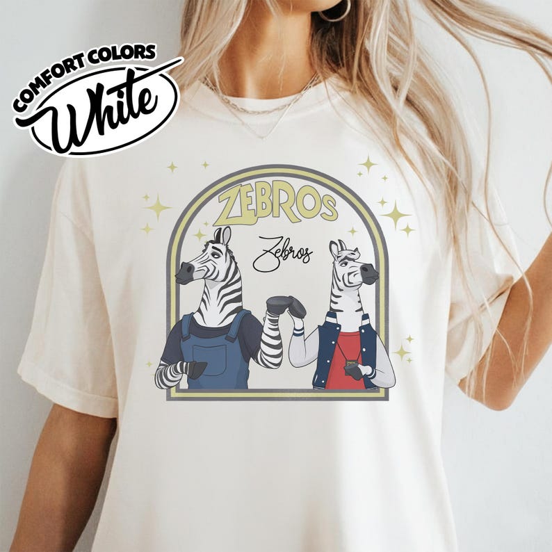 May include: Cream-coloured t-shirt with a graphic of two zebras in a stylized design. The zebras are depicted in a friendly pose, with the word "ZEBROS" and "Zebros" in a decorative font above them.