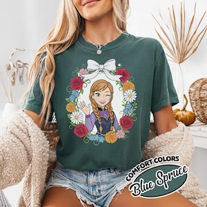 Peut inclure: T-shirt Comfort Colors Blue Spruce avec un personnage de dessin anim&eacute; dans une couronne florale. Le personnage a des tresses, fait un clin d'&oelig;il et salue. La couronne comprend des roses rouges, des marguerites et d'autres fleurs. Le t-shirt porte les mots "Comfort Colors" et "Blue Spruce".