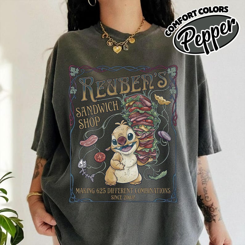K&ouml;nnte beinhalten: Dunkelgraues T-Shirt mit einer Comicfigur, die ein gro&szlig;es Sandwich h&auml;lt. Auf dem Shirt steht "Reuben's Sandwich Shop" und "Making 625 Different Combinations Since 2003!". Au&szlig;erdem ist der Text "Comfort Colors Pepper" aufgedruckt.
