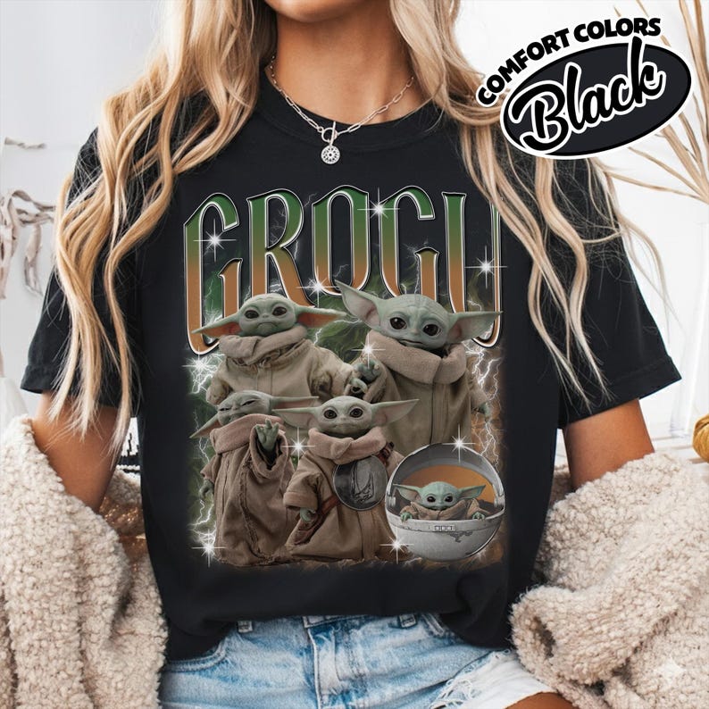 K&ouml;nnte beinhalten: Ein schwarzes T-Shirt mit einem Grafikdruck von mehreren Grogu-Figuren in beigen Roben. Der Text GROGU ist in gro&szlig;en, stilisierten gr&uuml;nen und orangefarbenen Buchstaben &uuml;ber den Figuren abgebildet. Das Design enth&auml;lt Blitzeffekte und Sterne.