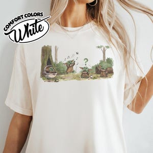 May include: White Comfort Colors t-shirt featuring a cartoon illustration of Grogu in a forest setting. The graphic shows Grogu in various poses, with musical notes, a question mark, and a small ball. The shirt has the words "Comfort Colors" and "White" in a circular design.