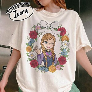 Peut inclure: T-shirt ivoire avec un personnage de dessin anim&eacute; aux tresses blondes, faisant un clin d'&oelig;il et saluant. Le personnage est entour&eacute; d'une couronne florale avec des roses rouges, des marguerites et des fleurs bleues. Le t-shirt porte les mots "COMFORT COLORS" et "Ivory".