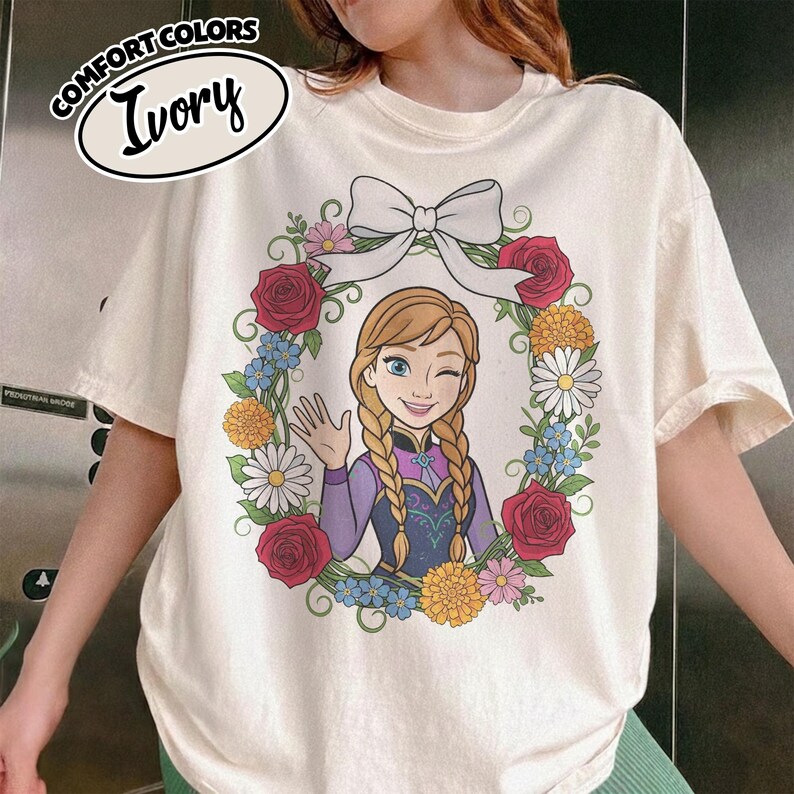 Peut inclure: T-shirt ivoire avec un personnage de dessin anim&eacute; aux tresses blondes, faisant un clin d'&oelig;il et saluant. Le personnage est entour&eacute; d'une couronne florale avec des roses rouges, des marguerites et des fleurs bleues. Le t-shirt porte les mots "COMFORT COLORS" et "Ivory".