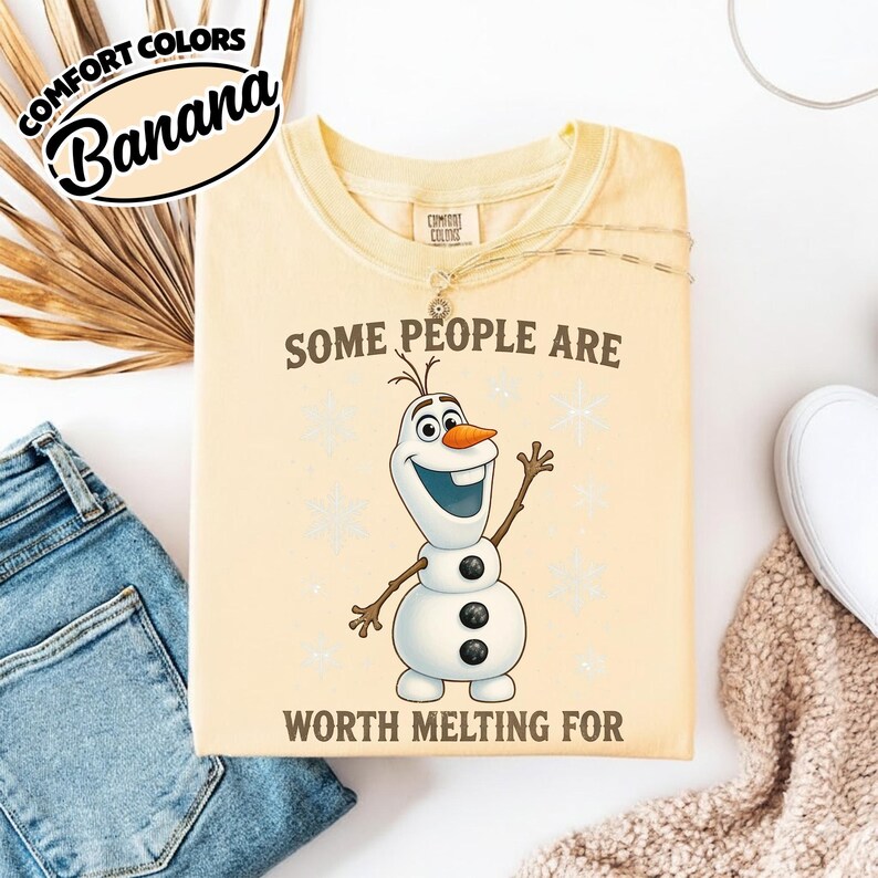May include: A light yellow t-shirt with the text "SOME PEOPLE ARE WORTH MELTING FOR" and an illustration of Olaf the snowman. The shirt also has the text "COMFORT COLORS" and "Banana" in a circular design.