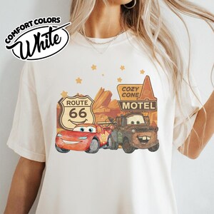 May include: White t-shirt featuring a graphic of Lightning McQueen and Mater from the movie Cars, with a Route 66 sign and Cozy Cone Motel. The shirt has the words "Comfort Colors" and "White" in a circular design.