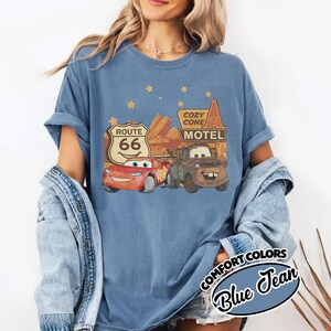 May include: Blue Comfort Colors t-shirt featuring a graphic of characters from the movie Cars, including Lightning McQueen and Mater, with Route 66 and Cozy Cone Motel signage. The shirt is a blue jean color.