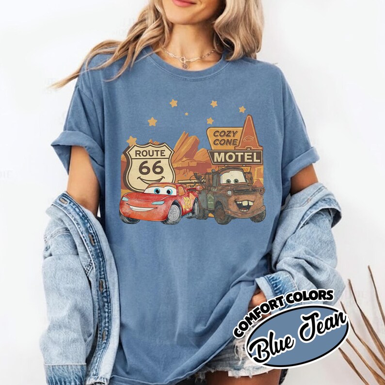May include: Blue Comfort Colors t-shirt featuring a graphic of characters from the movie Cars, including Lightning McQueen and Mater, with Route 66 and Cozy Cone Motel signage. The shirt is a blue jean color.