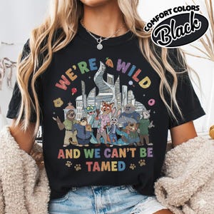 May include: Black t-shirt with a colorful graphic featuring characters from a cartoon movie. The text "WE'RE WILD AND WE CAN'T BE TAMED" is displayed in rainbow colors. The shirt also has the words "COMFORT COLORS" and "Black" in a circular design.